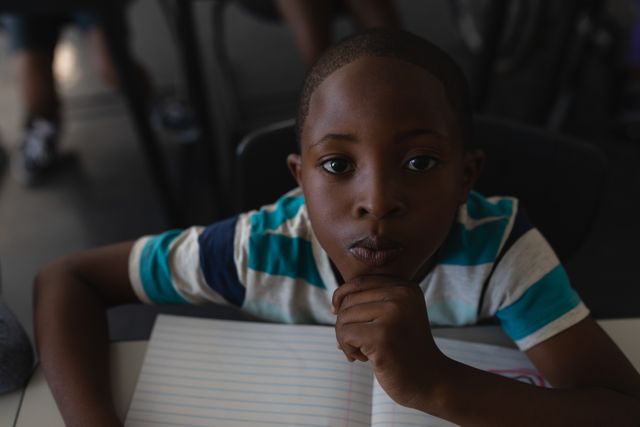 Young black schoolboy sitting at desk in classroom, hand on chin, looking at camera. Ideal for educational content, school promotions, childhood development articles, and academic materials.