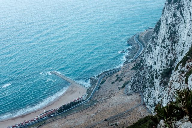 High-angle view capturing rugged coastal cliffs meeting the ocean. Winding, serpentine roads curve along the shoreline. Use in travel promotions, nature blogs, outdoor adventures content, road trip planning material.