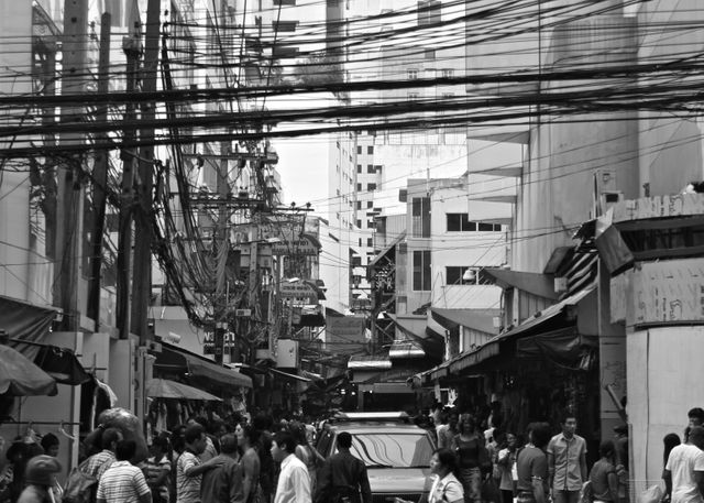 This black and white image depicts a busy urban street with crowded sidewalks, electric wires crisscrossing above, and high-rise buildings in the background. Ideal for illustrating urban life, bustling markets, and vibrant city atmospheres in articles, blogs, or websites focused on metropolitan areas and city living.