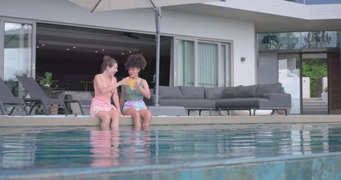Diverse women sitting on pool edge clinking yellow cups under umbrella at modern villa