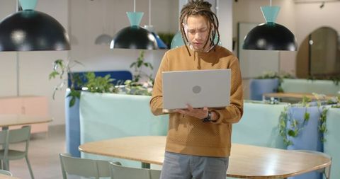 African American man working on laptop standing in modern coworking lounge under pendant lamps
