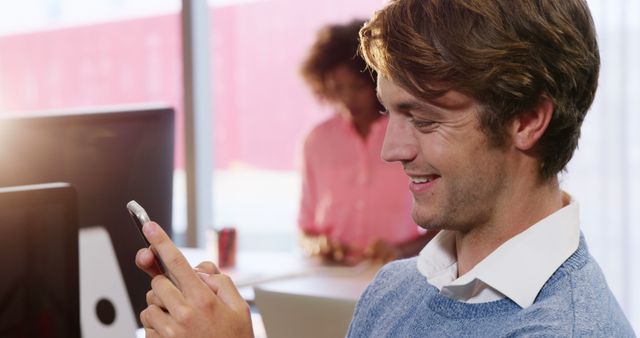 Young professional man happily using smartphone in a modern office setting. He is seated at a desk while casually looking at his screen. This image can be used to illustrate themes of workplace communication, modern technology use, and casual work environments. Ideal for promoting tech devices, communication platforms, or illustrating modern work culture.