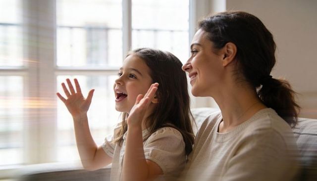 Mother and daughter sharing joy on couch by sunny window, daughter singing and waving