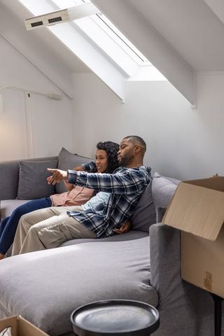 Diverse couple sitting on gray sectional in sunlit loft unpacking moving boxes, relaxing