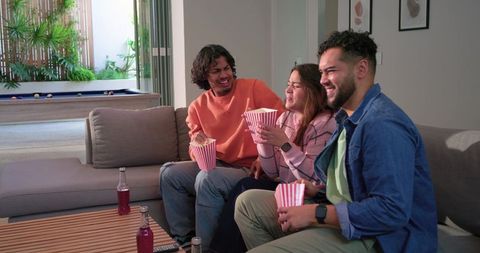 Diverse friends laughing and sharing popcorn on gray sofa in cozy modern living room