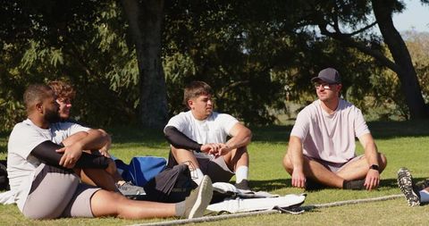 Multiracial male teammates sitting on grass, resting and chatting after outdoor training