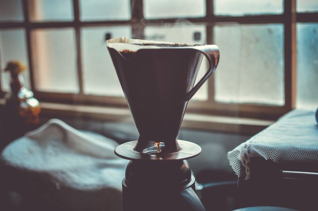 A rustic coffee dripper is sitting on top of a coffee container, basking in the soft morning light that streams through a slightly foggy window. The scene conveys a cozy and serene atmosphere, perfect for representing home coffee brewing, morning routines, or artisanal coffee traditions. Ideal for use in blogs, social media posts, and advertisements related to coffee, kitchen appliances, and lifestyle content.