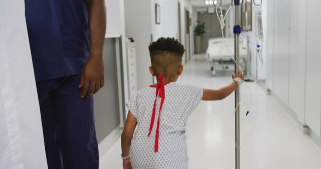 A young child patient is walking through a hospital corridor while holding onto an IV stand. He is wearing a hospital gown with a red tie. An adult in scrubs is walking beside him, suggesting medical care and supervision. This image can be used for depicting pediatric healthcare, hospital care, medical treatments, recovery stories, and health-related educational materials.