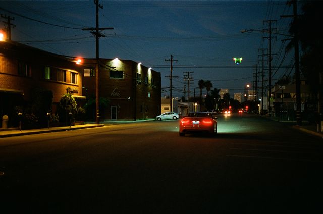 Red car driving through an empty city street at night. Streetlights illuminate buildings and sidewalk with a dim glow. Ideal for urban lifestyle, travel blogs, cityscape series, nighttime drives concepts.