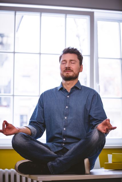 Young male executive practicing meditation in lotus position at a modern office. Ideal for illustrating concepts of workplace wellness, stress relief, mindfulness, and work-life balance. Suitable for articles, blogs, and promotional materials related to mental health, corporate wellness programs, and relaxation techniques.