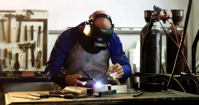 Welder wearing helmet and equipment welding metal pieces on workbench in workshop. Tools hanging on wall in background. Useful for articles about skilled labor, safety in the workplace, industrial professions, and craftsmanship.