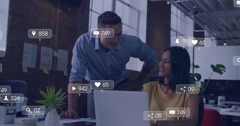 Coworkers collaborating over laptop with social media engagement metrics in modern office
