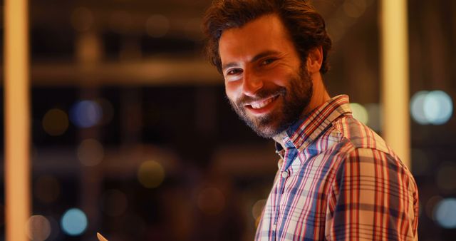 Man in plaid shirt smiling while holding tablet in modern office at night with blurred lights in background. Perfect for articles on modern work environments, business technology, or employee well-being. Great for advertising tech devices or professional lifestyle content.