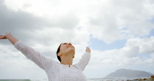 Woman stands with arms outstretched, embracing freedom and fresh air under cloudy sky by the mountains. Perfect for themes on wellness, mental health, outdoor activities, and finding inner peace.