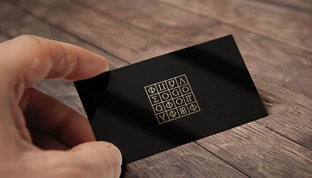 Hand holding black foil business card with gold geometric glyph grid on wood tabletop