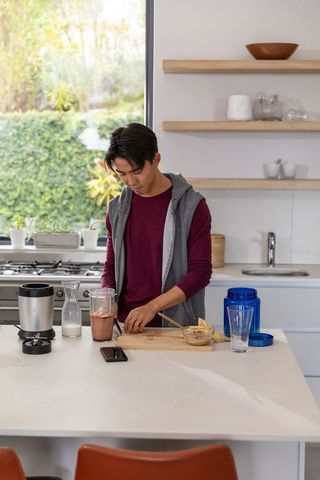 Asian man preparing banana smoothie at modern kitchen island with blender and smartphone
