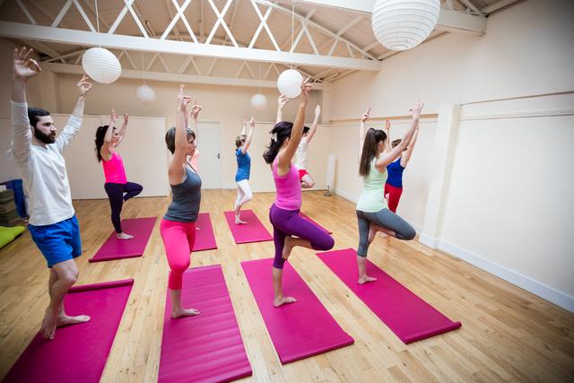 Group of individuals practicing yoga together in a modern fitness studio. They are performing a yoga pose on pink mats, focusing on balance and mindfulness. Ideal for use in articles or advertisements related to fitness, wellness, group activities, and healthy lifestyles.