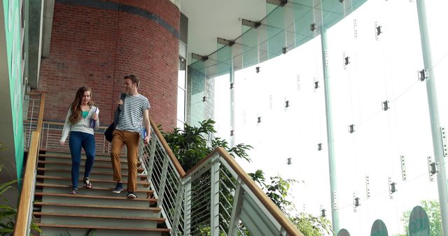 Two young people walk down stairs in a modern building with large windows and ample natural light. Both are casually dressed, suggesting a relaxed and informal atmosphere. Suitable for content related to university life, young professionals, or modern architecture.