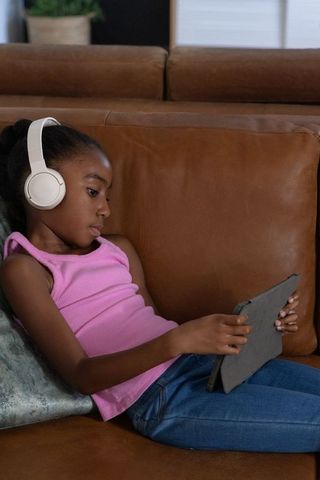 African american girl reclining on leather sofa with headphones using tablet for learning
