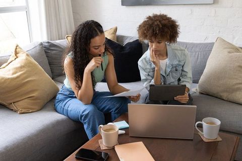 African american women collaborating on laptop and tablet while reviewing documents at home