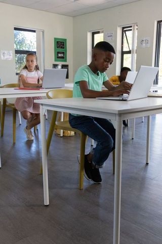 Elementary students using laptops in bright classroom, boy focusing on screen learning