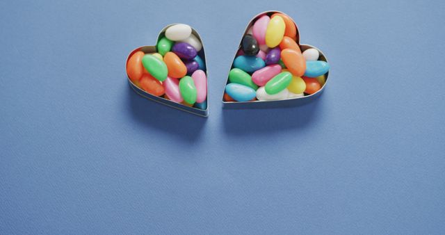 Colorful jelly beans presented in heart-shaped cookie cutters are perfect for conveying themes of love and sweetness. This photo can be ideal for use in Valentine's Day promotions, candy advertisements, or any content related to desserts and sweets. The vibrant colors against a blue background create a lively and appealing visual, suitable for posters, social media banners, and holiday greeting cards.