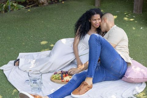 Couple reclining on picnic blanket sharing fresh fruit board in sunlit backyard romance