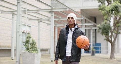 African american man walking with basketball wearing beanie and headphones outdoor campus