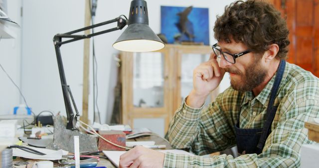 This image shows a focused artist sitting at a cluttered desk in his workshop, thoughtfully sketching under a lamp. The workspace is filled with various tools and materials, indicating a creative and productive environment. Ideal for articles or websites related to artistry, craftsmanship, creative processes, or hobbies.