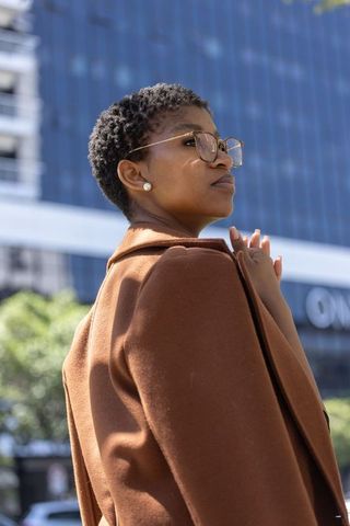 Confident african woman standing in city wearing brown coat and glasses, chic professional look