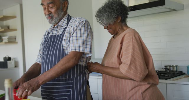 Elderly Black couple enjoying precious moments in a modern kitchen while cooking. The couple wears aprons, highlighting domestic life and co-operative household activities. Ideal for content relating to family values, healthy lifestyles, and senior relationships. Can be used in articles, blog posts, or marketing campaigns promoting togetherness, senior wellness, or cooking.