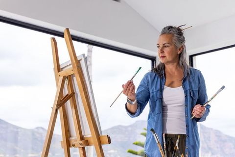 Mature woman painting in mountain-view studio, holding brushes and checking brushstroke