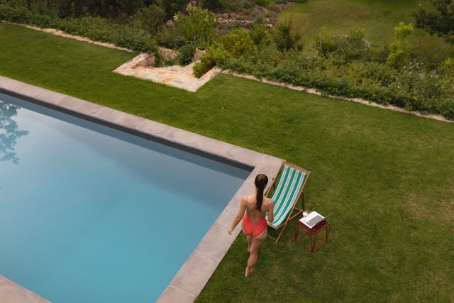 High angle view of a woman in swimwear standing near the poolside in a backyard. Ideal for use in advertisements for summer vacations, outdoor activities, relaxation products, or lifestyle blogs. The serene and tranquil setting makes it suitable for promoting wellness and leisure.