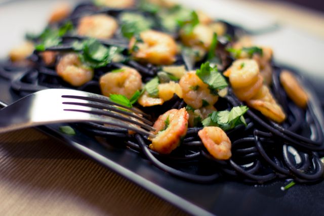 Close-up view of squid ink pasta served with shrimp and garnished with fresh herbs, showcasing vibrant colors and textures. Useful for food blogs, restaurant menus, culinary articles, or advertisements promoting gourmet dining experiences.