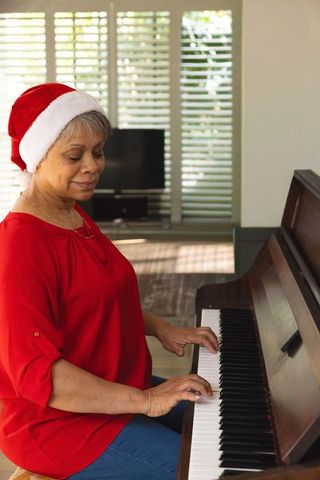 Mature african american woman playing piano at home wearing santa hat for christmas