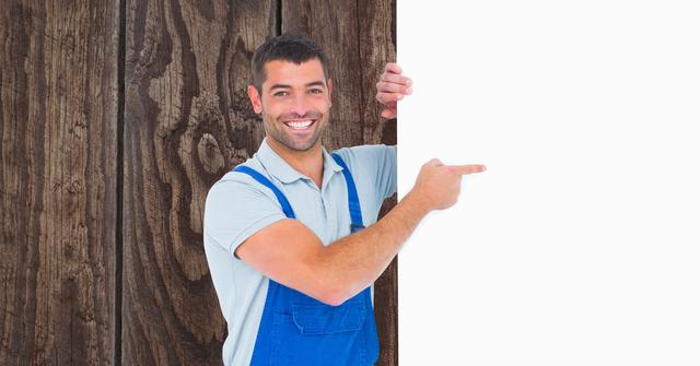 Smiling male worker in blue overalls pointing at a blank billboard against a wooden background. Ideal for advertisements, promotional materials, marketing campaigns, and construction or handyman services. The blank billboard provides ample space for custom messages or branding.