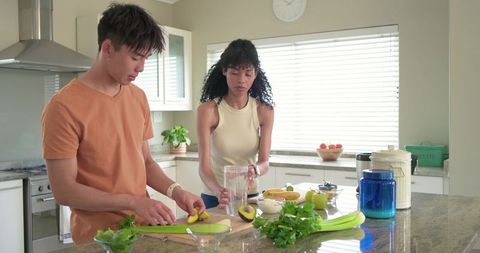 Diverse couple preparing avocado smoothie on bright modern kitchen island with fresh herbs