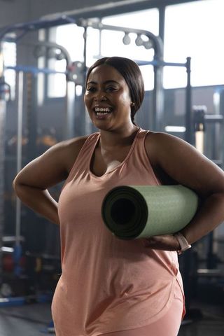 African american woman holding yoga mat in gym smiling in pink activewear