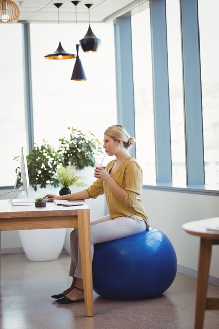 Businesswoman sitting on a fitness ball while working at her desk in a modern office. The scene highlights a healthy lifestyle and ergonomic workspace. Ideal for use in articles or advertisements about workplace wellness, productivity, modern office design, and ergonomic office solutions.
