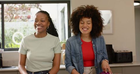 Female friends preparing fresh salad in bright kitchen, smiling and laughing together