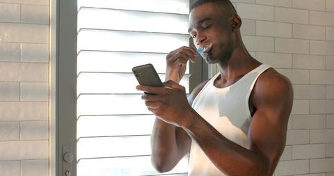 African American Man Brushing Teeth While Using Smartphone in Sunlit Minimal Bathroom