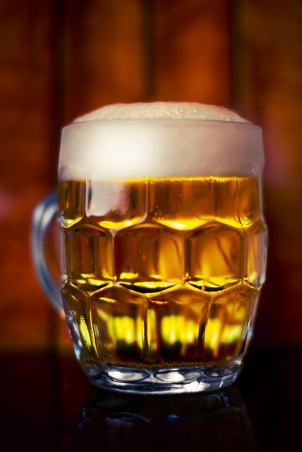 A glass mug filled with golden beer, topped with a thick layer of foam, positioned against a warm wooden backdrop. Ideal for use in advertisements, menus, articles on brews and pubs, marketing materials for bars and restaurants, and social media posts related to food and drink.