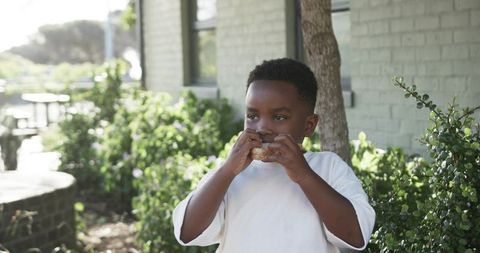 African preteen boy eating sandwich outdoors by painted brick wall with green shrubs