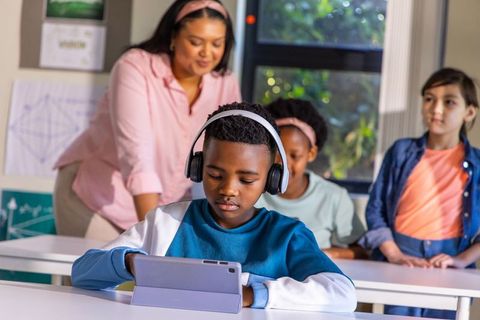 Elementary student using tablet with headphones while teacher guiding classroom learning