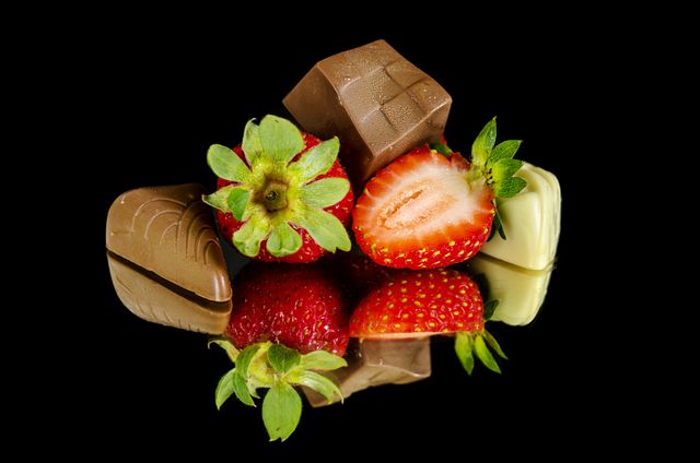 Stylized arrangement of assorted chocolates and whole and sliced strawberries presented against black background. Ideal for use in marketing materials for chocolatiers, dessert recipes, restaurant menus, Valentine's Day promotions, or gourmet food blogs.