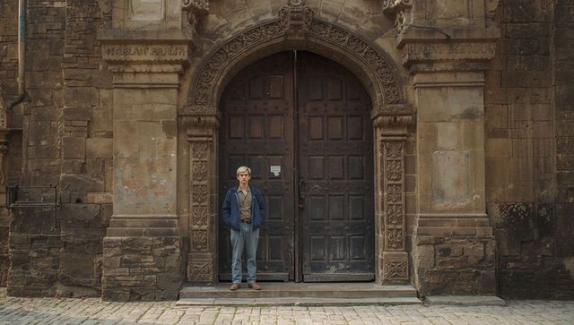 Young man standing before ornate arched wooden doors of historic weathered stone facade