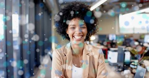 Smiling professional woman standing in sunlit open-plan office wearing beige blazer, bokeh
