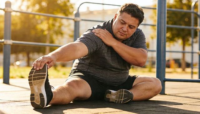 Overweight man stretching leg in sunlit urban park preparing for outdoor workout