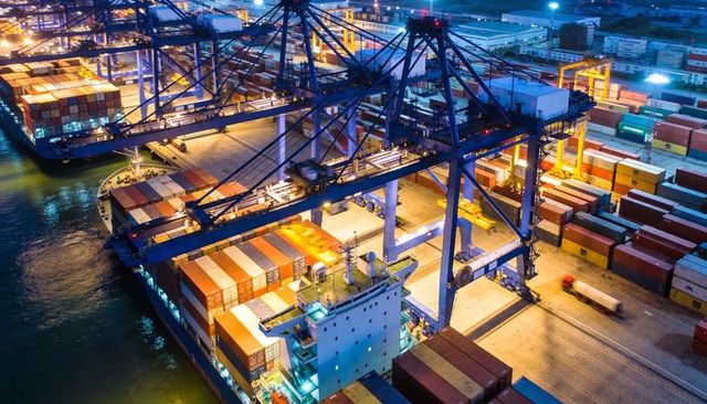 Night port cranes loading containers onto berthing cargo ship under floodlights