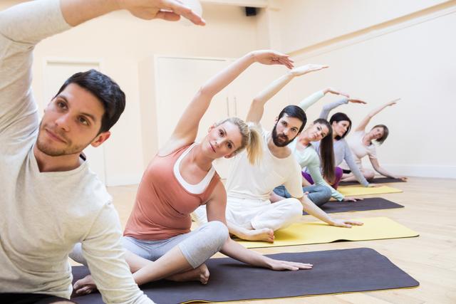 Group of adults performing yoga stretches in a fitness studio. Everyone is aligned in a row on yoga mats, concentrating on their flexibility and balance. Ideal for illustrating concepts of group exercise, healthy living, and wellness programs. Can be used in fitness blogs, gym promotions, or lifestyle magazines.